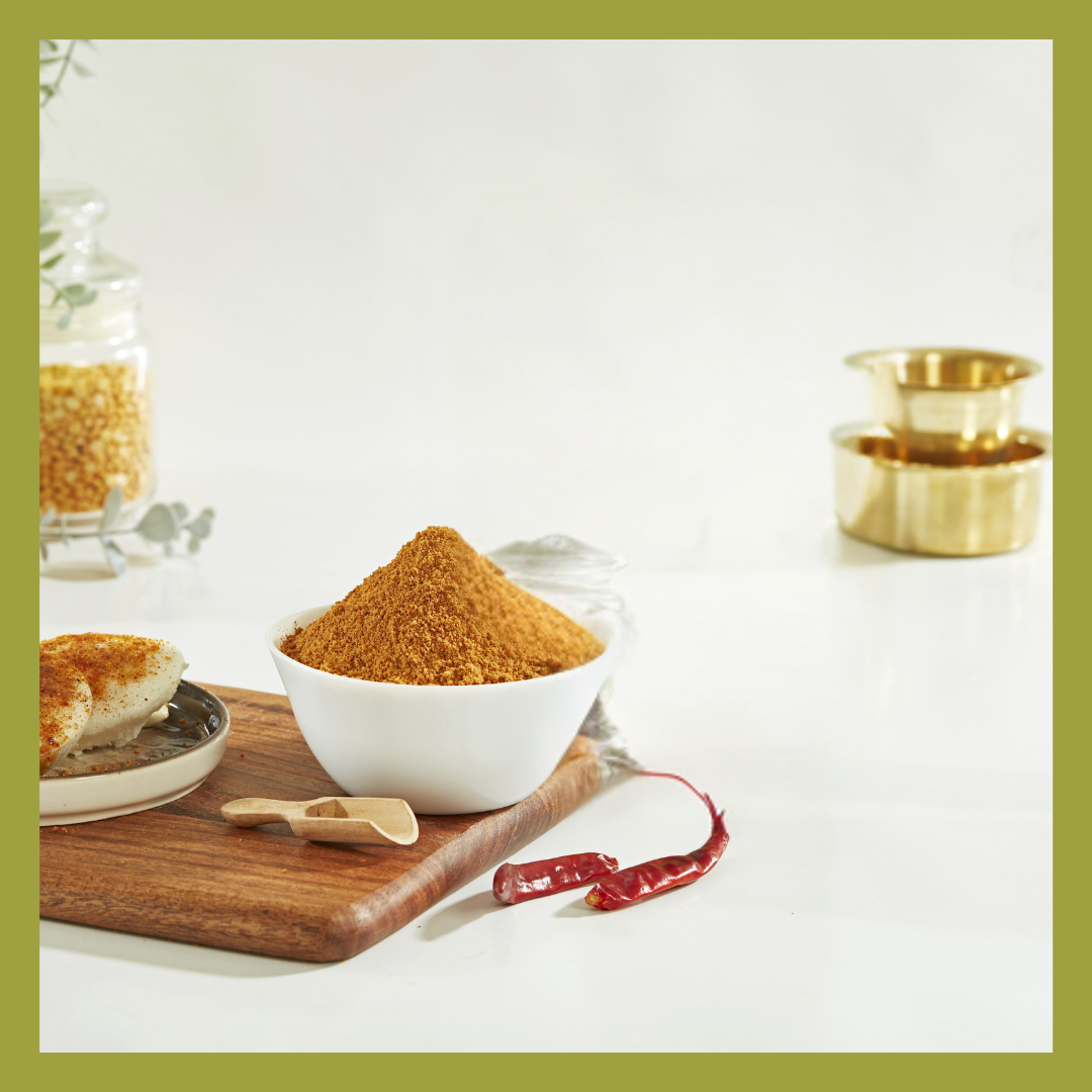 White bowl filled with Malu's Idly dosa molaga podi spice powder on wooden board with red chili and idly
