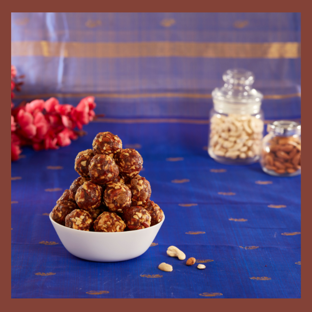 Malu's South Indian sweets dry fruit ladoo stacked in a white bowl on a blue cloth with nuts jars in the background